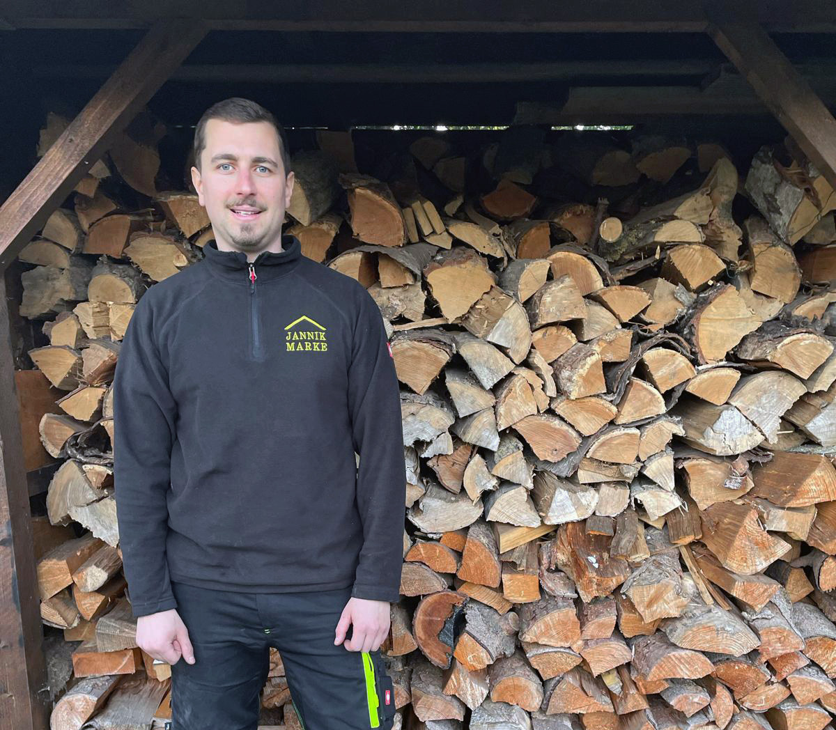 Janik Marke in Arbeitskleidung vor Brennholzstapel